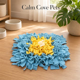 A square pet snuffle mat, featuring a blue outer layer and a yellow inner layer, displayed on a wooden floor in a bright room with a plant and bowls.