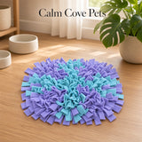A square pet snuffle mat made of blue and purple felt strips, arranged in a blue plus sign with purple corners on a light wood floor. The background shows a cozy room setting with a potted monstera plant, ceramic bowls, and a window, with the text 'Calm Cove Pets' at the top.