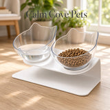 A white dual pet elevated feeder and waterer bowl set with clear glass cat-shaped bowls, one filled with water and the other with dry cat food, placed on a light wood floor in a bright room. The text 'Calm Cove Pets' is overlaid at the top.