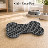 A gray bone-shaped silicone mat with a honeycomb texture, placed on a light wood floor in a home setting. Two pet bowls are visible in the background, and the text 'Calm Cove Pets' is displayed at the top.