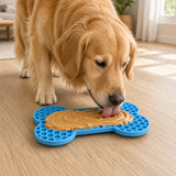 A golden retriever dog licking a bone-shaped blue silicone treat mat filled with peanut butter on a light wood floor in a bright room.