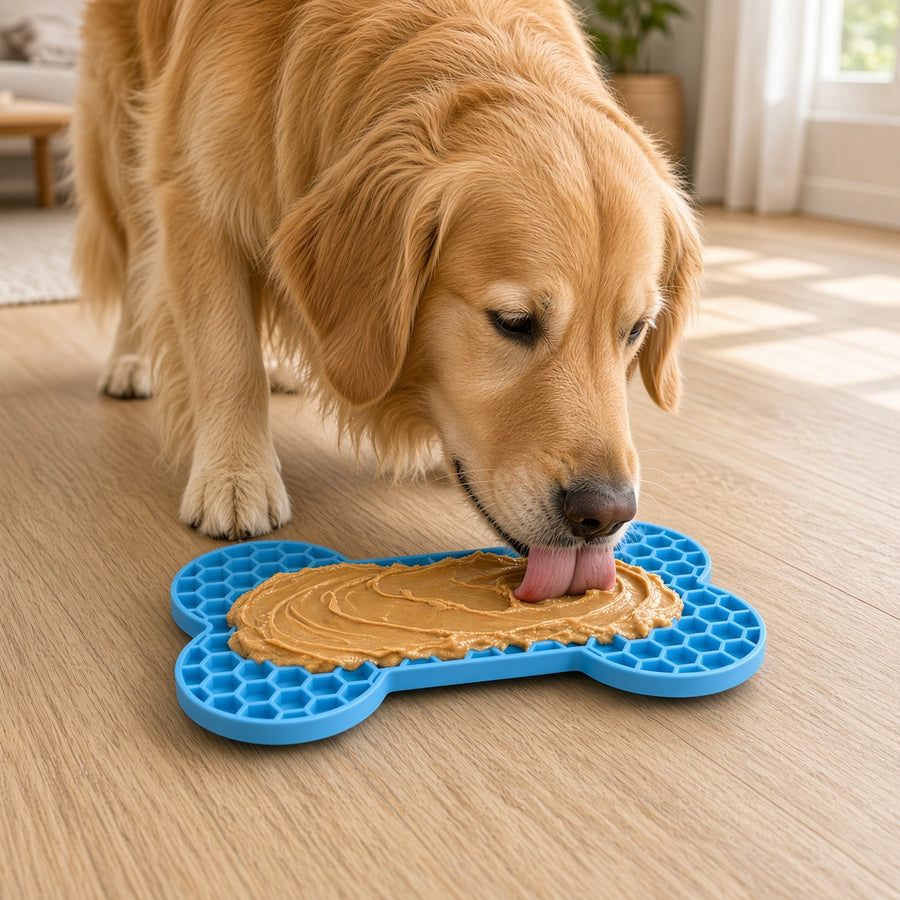 A golden retriever dog licking a bone-shaped blue silicone treat mat filled with peanut butter on a light wood floor in a bright room.