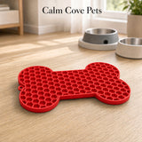 A bright red bone-shaped silicone mat with a honeycomb texture, placed on a light wood floor in a home setting. Two pet bowls are visible in the background, and the text 'Calm Cove Pets' is displayed at the top.