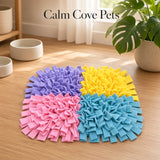 A four-colored felt pet snuffle mat on a wooden floor, with the text 'Calm Cove Pets' above it. The mat is divided into four quadrants in purple, yellow, pink, and blue, with small white bowls and a potted plant in the background.