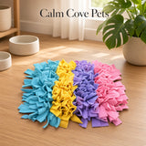 A pet snuffle mat with four vertical color panels in blue, yellow, purple, and pink, displayed on a light wood floor in a bright room with a plant and bowls in the background. The text 'Calm Cove Pets' is visible at the top.
