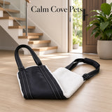 A black and white pet mobility support sling with a white sherpa lining, placed on a light wood floor in a modern home interior with a staircase and potted plant in the background. The text 'Calm Cove Pets' is displayed at the top.