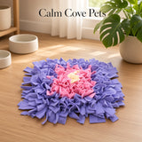 A square pet snuffle mat with a concentric design in purple, pink, and cream, displayed on a light wood floor in a bright room with a plant and bowls in the background. The text 'Calm Cove Pets' is visible at the top.