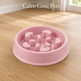 A pink slow feeder pet bowl with raised textured nubs inside, placed on a light wood floor near a window. The text 'Calm Cove Pets' appears at the top.
