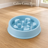 A light blue slow-feeding pet bowl with raised textured nubs inside, shown on a wood floor in a bright room. The text 'Calm Cove Pets' appears near the top.