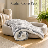 A gray and white patterned pet anxiety blanket is draped over a beige pet bed in a bright, sunlit living room. The text 'Calm Cove Pets' is displayed at the top of the image.