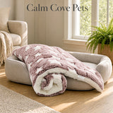 A rolled plush pink pet anxiety blanket with a dusty mauve and cream star pattern draped over a beige pet bed in a bright, sunlit living room. The text 'Calm Cove Pets' is displayed at the top.