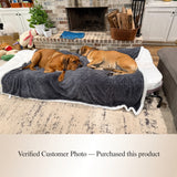 Two dogs lying on a large pet lounger with a dark gray plush weight anxiety blanket and white trim in a living room. A text banner at the bottom reads 'Verified Customer Photo — Purchased this product'.