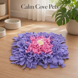 A video of a square pet snuffle mat with a concentric design in purple, pink, and cream, displayed on a light wood floor in a bright room with a plant and bowls in the background. The text 'Calm Cove Pets' is visible at the top.